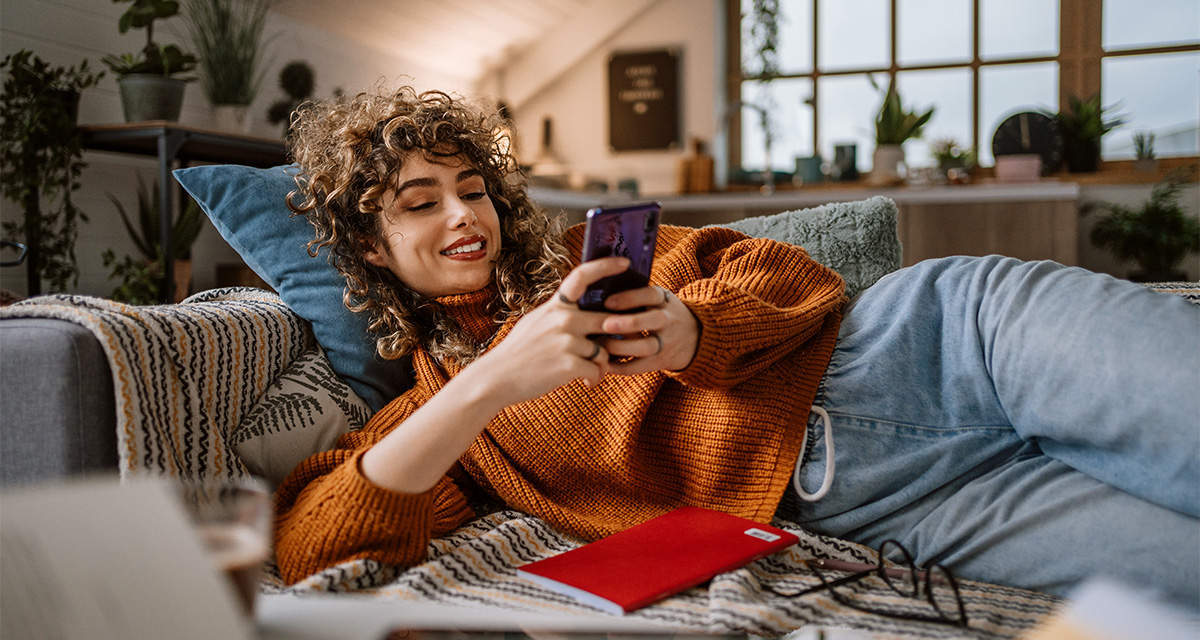 Una ragazza legge rilassata lo smartphone su un divano: risparmiare sul riscaldamento vuol dire qualità della vita
